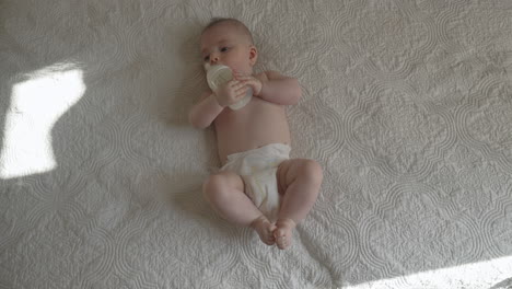 Newborn-trying-self-feeding-with-milk-bottle-playful-mood-in-white-sunny-bedroom,-happy-baby