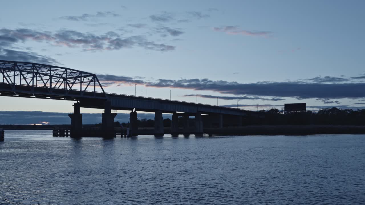 Semi-truck drives across a long bridge at dusk under dramatic blue clouds. Reflections shimmer in the river below