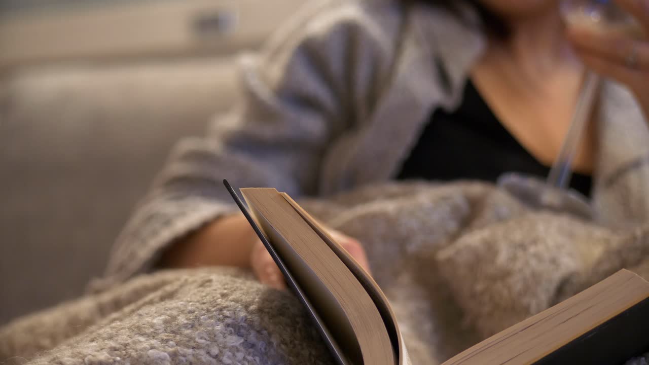 居心地の良いヒュッゲのコンセプト - ベージュのブランケットにくるまりリビングのソファで本を読む女性、スライダーによるクローズアップショット