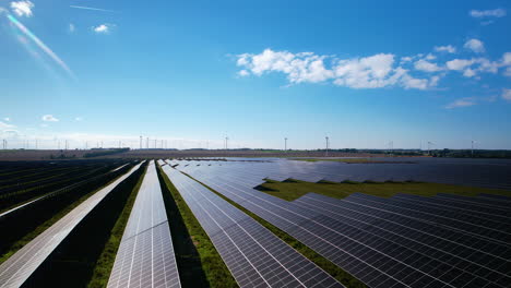 Luftdrohne,-Die-An-Sonnenkollektoren-Mit-Windmühlen-Im-Hintergrund-Vorbeifliegt