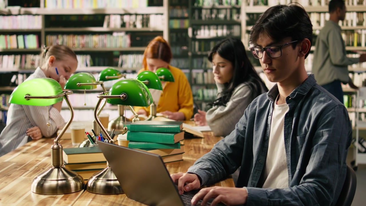 Studente asiatico lavora al laptop in una biblioteca tranquilla con alti scaffali di libri.