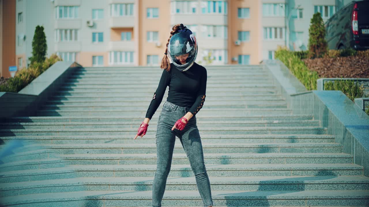 pretty woman in gray jeans and with helmet on her head is posing on the background of a red motorbike and urban high-rise buildings