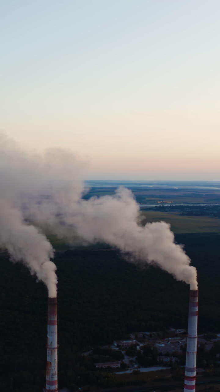 White smoke from pipes on nature background. Two chimneys produce emissions into the air among green fields and lakes. Vertical video