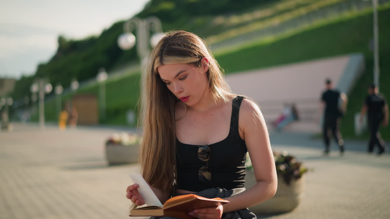 Woman seated outdoors on a bench with legs crossed, flipping a page from her book, thoughtfully observing a piece of paper, and placing it back, the background, people walking, and distant lampposts