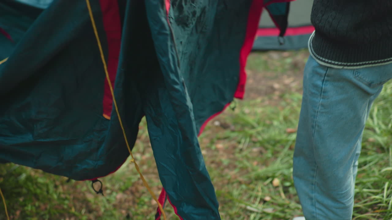 Camper crouches on grassy forest ground and lifts tent fabric while inspecting material during camping setup, with hands holding edge of tarp and ropes spread across surface for alignment