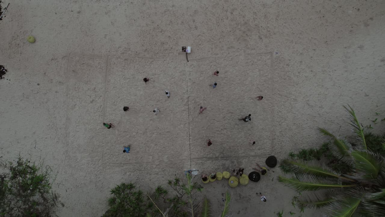 Top-down aerial view of children playing football on an improvised sandy beach playground, surrounded by palm trees in a serene tropical setting. Location Philippines.