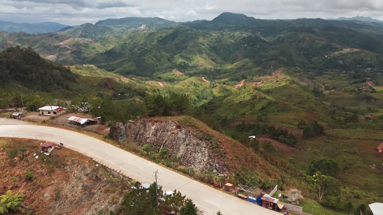 Scenic 4K aerial drone flight over a paved two-lane road winding along a steep mountain ridge. Panoramic view of rolling green hills, rural settlements, and distant valleys