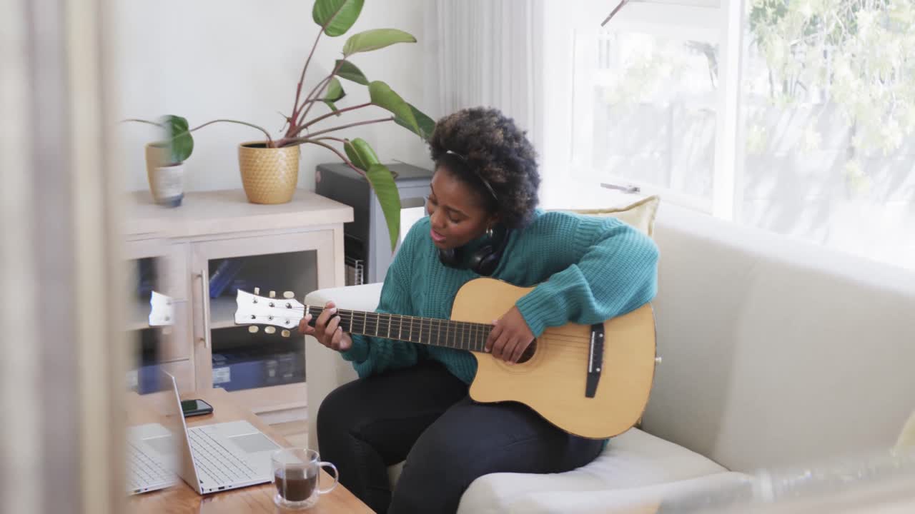 Donna afroamericana felice che si esercita a suonare la chitarra usando un laptop a casa, al rallentatore.