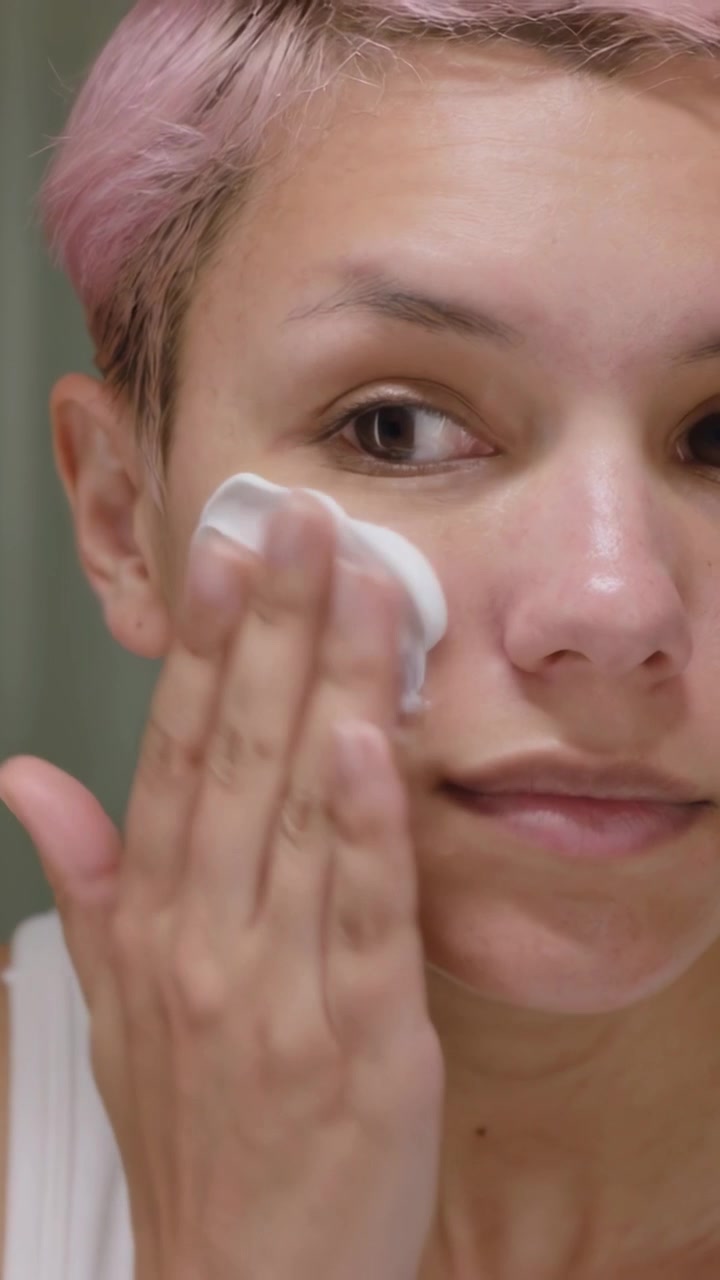 Vertical video: Pressing pad she with pink hair in white tank rubbing cream at vanity for skincare
