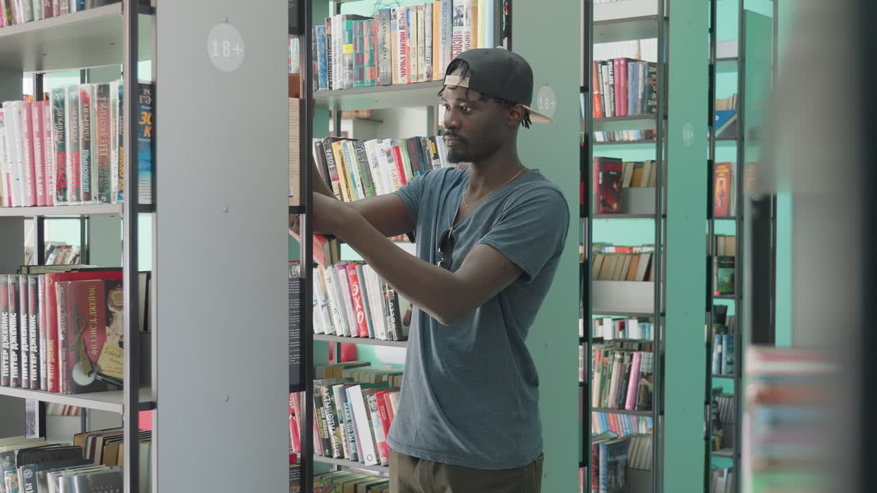 Young college man in casual clothing standing between library shelves, reaching for book with focused expression, surrounded by neatly arranged colorful spines in bright, modern interior