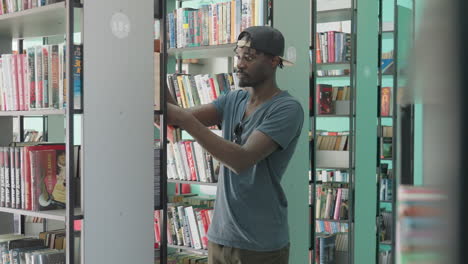 young college man in casual clothing standing between library shelves, reaching for book with focused expression, surrounded by neatly arranged colorful spines in bright, modern interior