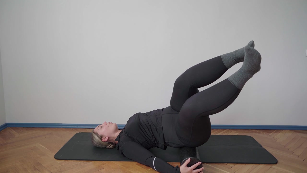 Adult exerciser performs lying leg raises while balanced on a foam roller to train core control and support the lumbar spine.