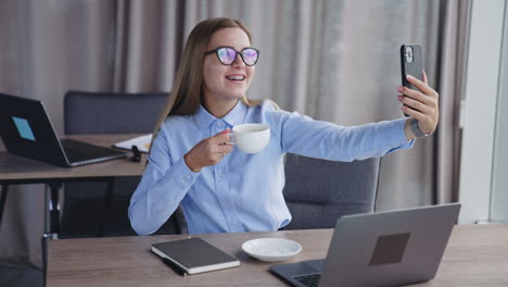 young blonde smiling lady having coffee sitting at table. woman taking selfie with a cup on her phone.