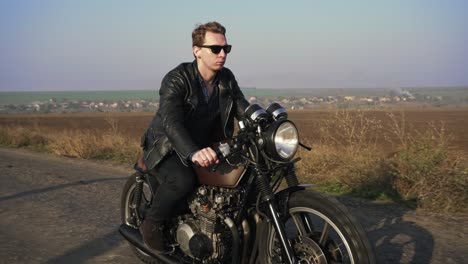 Side-view-of-a-stylish-cool-young-man-in-sunglasses-and-leather-jacket-driving-his-chopper-on-a-asphalt-road-on-a-sunny-day.-Shot-in-4k