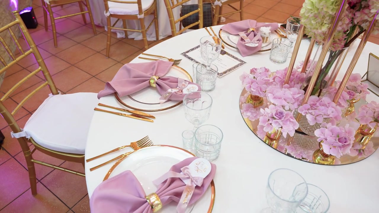 Stylish wedding reception table set with gold cutlery, blush napkins and glassware, featuring a mirrored centerpiece with pink flowers under soft, ambient lighting for a romantic mood.