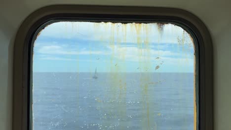 sailboat sailing on calm sea as seen through dirty and rusty window of navigating old ferry