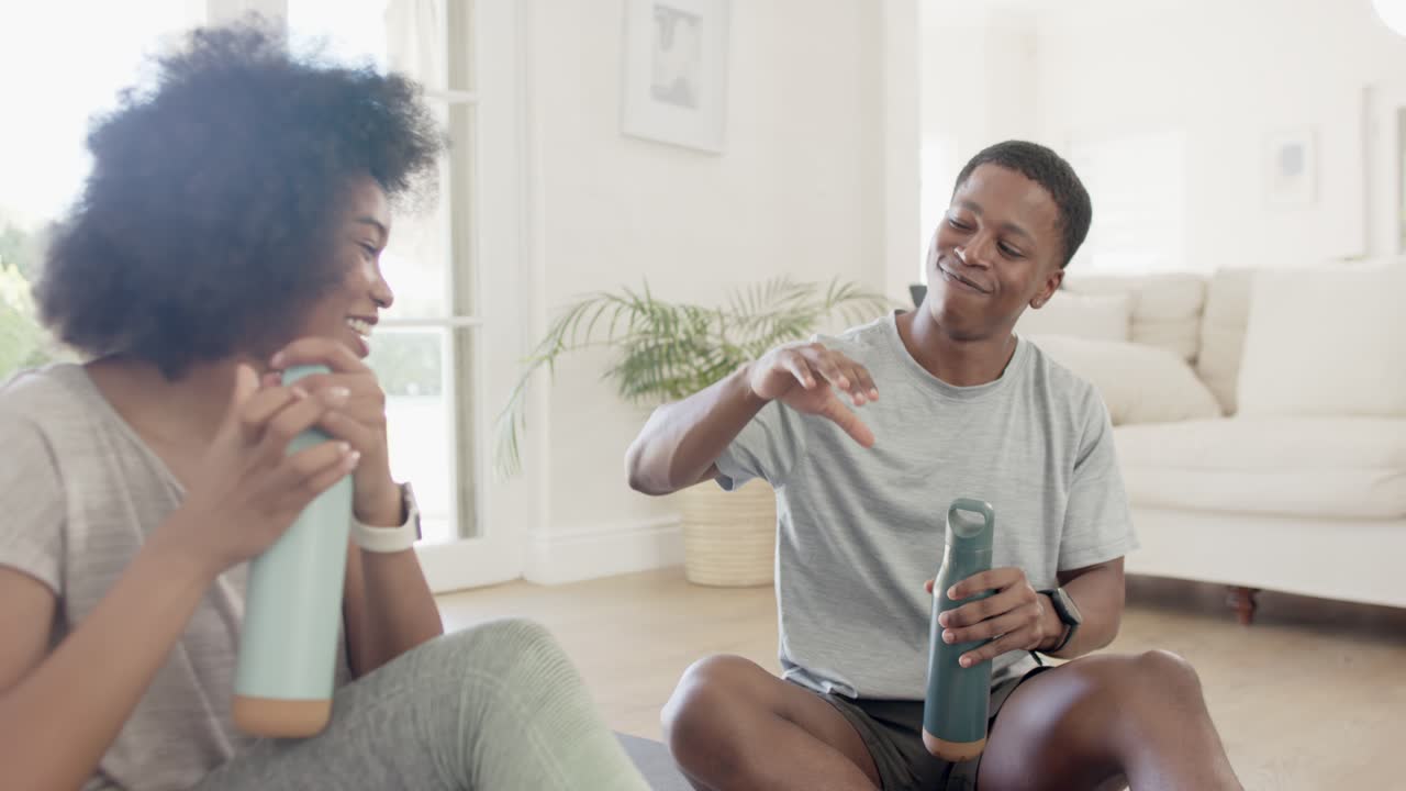 Pareja afroamericana sentada en colchonetas de yoga en casa, el hombre ofreciendo una botella, interactuando con alegría y sonrisas