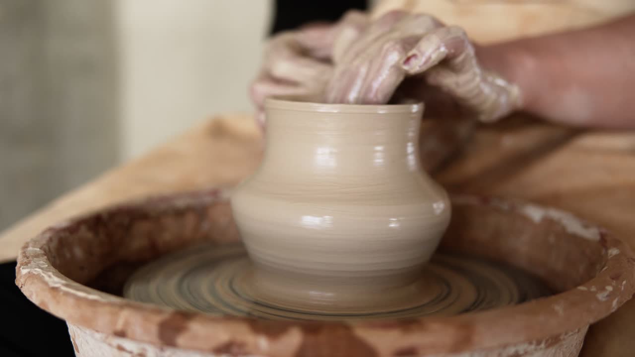 Close-up of potter's dirty hands working with wet clay on a pottery wheel making a vase in a workshop. Unrecognizable female person forming product