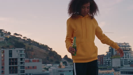 beautiful young african american woman with stylish afro walking on rooftop edge enjoying weekend lifestyle drinking alcohol at sunset