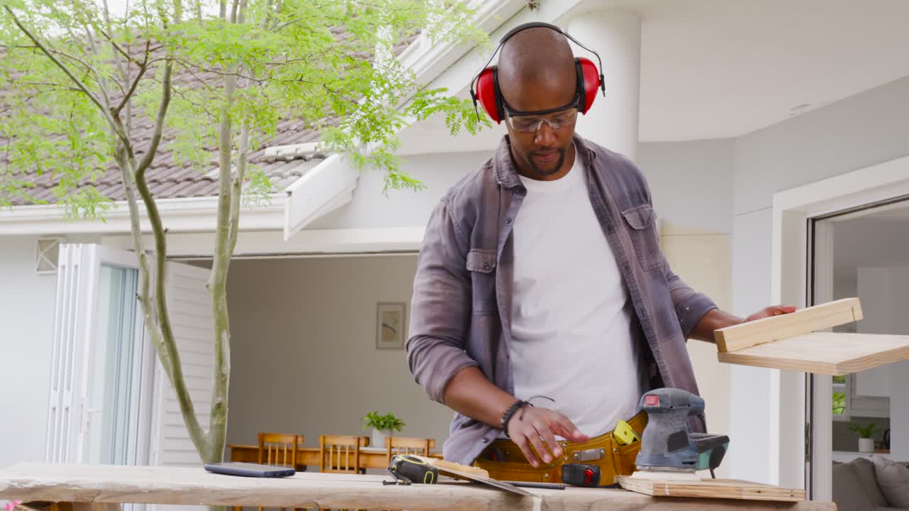 Man checking boards then sanding on patio with orbital sander, with earmuffs smoothing wood