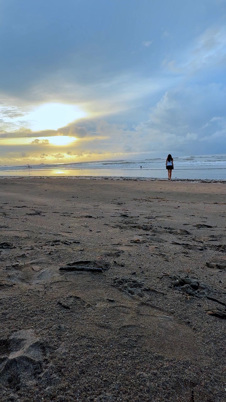 Pessoas caminhando em direção à arrebentação do oceano ao pôr do sol na Costa Rica. Vista de baixo ângulo mostra pegadas na areia molhada sob nuvens em camadas e luz amarela refletida perto do horizonte