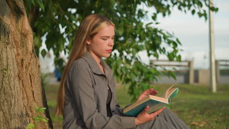 lady seated outdoors flipping pages of book in hand, looking thoughtful, greenery and blurred view of people playing in background, sunlight softly illuminates face as she contemplates