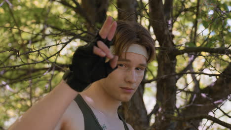 young soldier in green tank top stands beneath leafy branches in dense forest raising two fingers in friendly hi gesture playfully while winking at camera capturing adventurous spirit