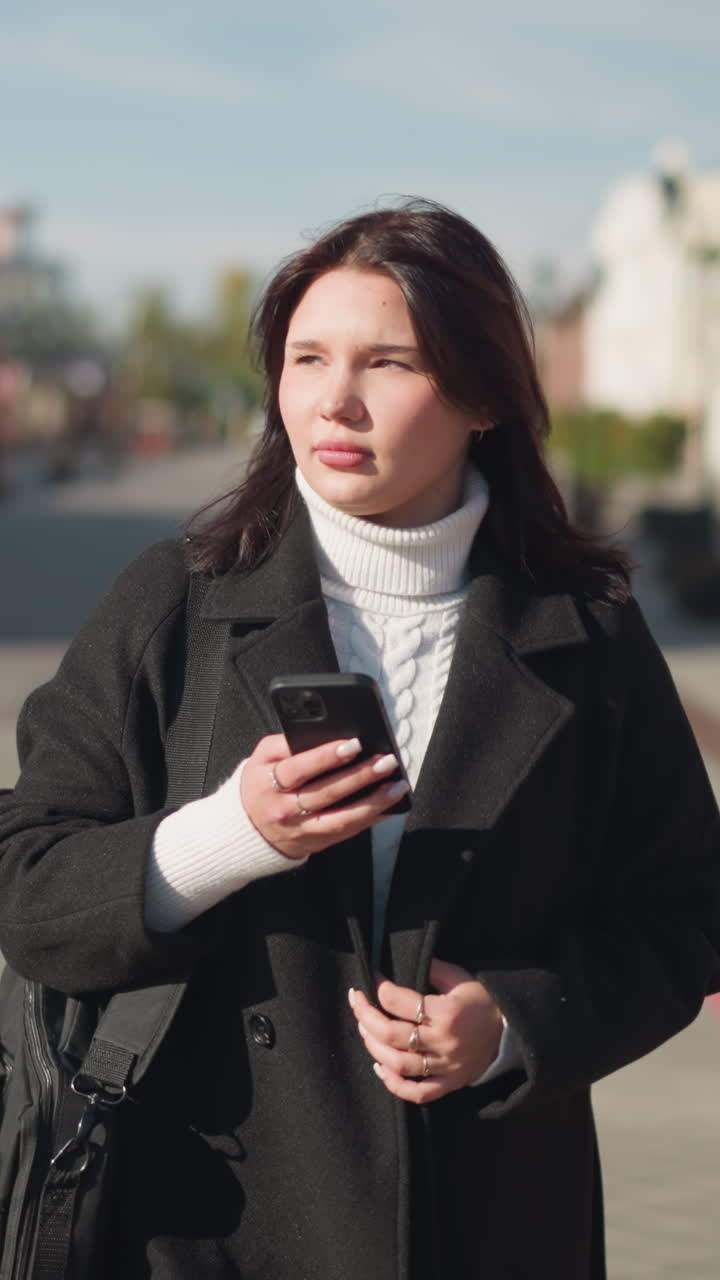 стильная женщина в черном пальто и белом свитере идет по солнечной городской улице, сосредоточенная на своем смартфоне, на заднем плане очаровательная архитектура, размытая зелень и асфальтированная пешеходная дорожка