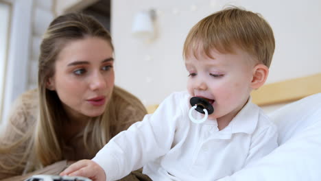 adorable caucasian baby boy with pacifier sits in the pillows comfortably. mother lying next to her son talks to him lovingly.