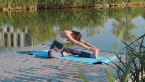 Fitte,-Kaukasische-Junge-Frau-In-Sportkleidung-Macht-Stretching-Übungen-Auf-Einer-Yogamatte-In-Der-Nähe-Eines-Teichs-Im-Park