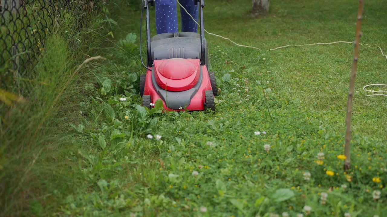 a woman is mowing grass and weeds with a lawn mower near a fence in the yard