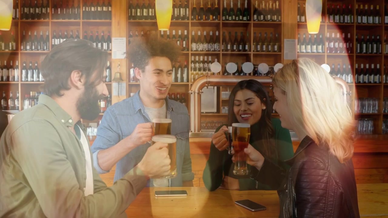 Uomo barbuto che offre un boccale di birra in una birreria, amici che brindano facendo tintinnare i bicchieri vicino a bottiglie e telefono.