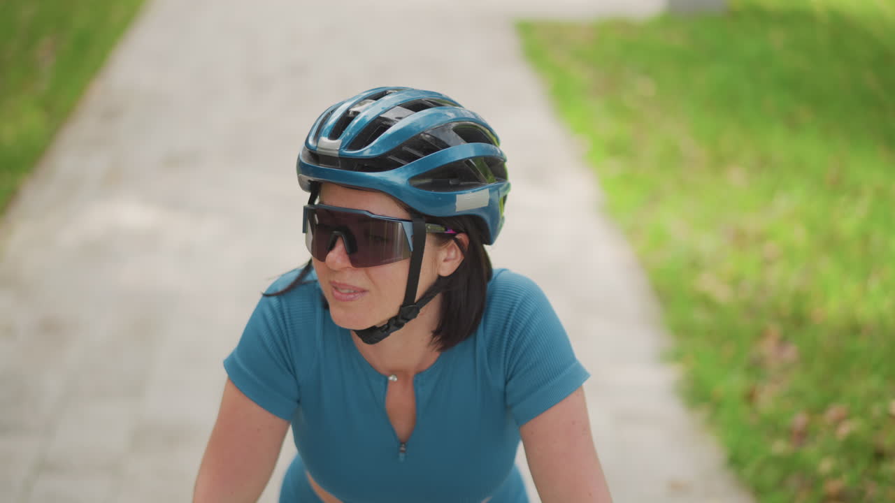 woman cycling on sunlit park path wearing blue top and helmet, mirrored sunglasses and focused expression, closeup front angle, steady cadence, leafy sidewalk, weekday training vibe, determined