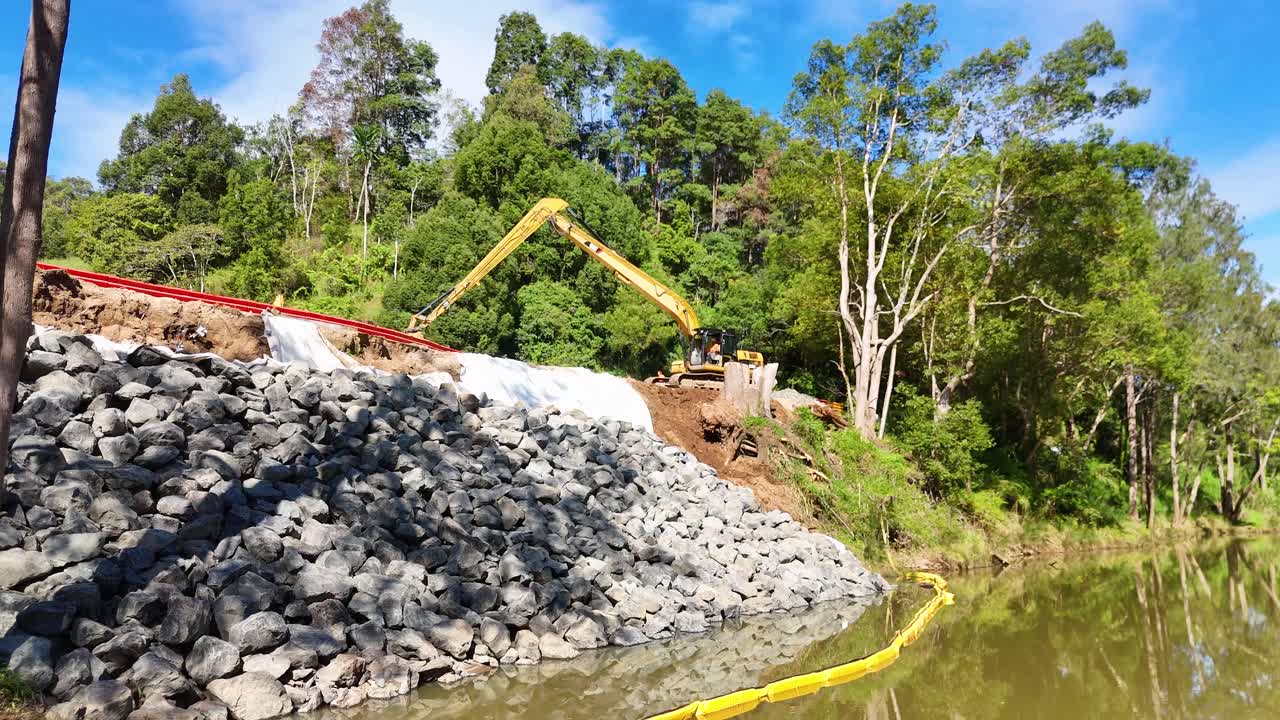Aerial view of construction equipment along a creek in Uki, NSW, under bright daylight. The scene includes lush greenery and a rocky embankment