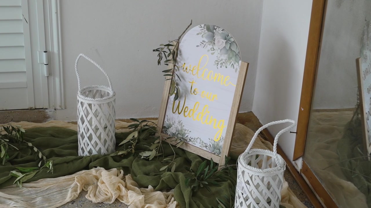 Et velkomstskilt er utstilt for et bryllup. Det har et blomsterdesign med ordene «Velkommen til vårt bryllup». Grønn og hvit dekor er arrangert rundt det. Kurver inneholder grønne planter