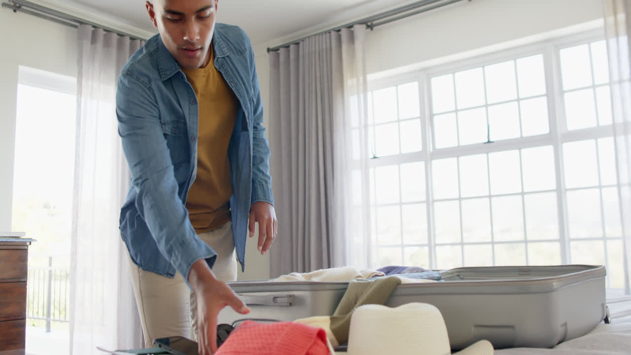 Happy biracial man packing suitcase in bright bedroom