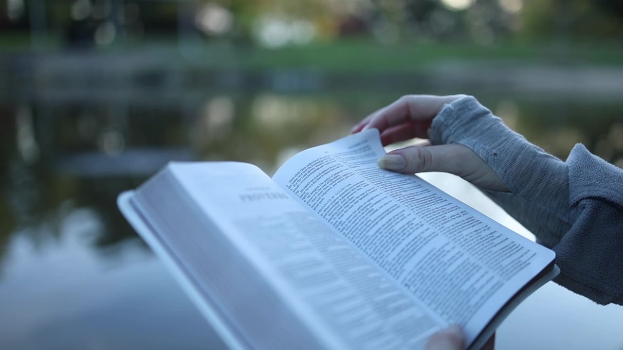 Primo piano di mani che sfogliano pagine in un libro aperto accanto a un lago tranquillo, luce naturale del giorno, profondità di campo ridotta, atmosfera rilassata, lettura all'aperto, atmosfera serena e riflessiva