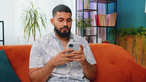Indisch-arabischer-Junger-Mann-Sitzt-Auf-Dem-Sofa-Und-Nutzt-Sein-Smartphone,-Um-Nachrichten-In-Einer-Social-Media-Anwendung-Zu-Teilen