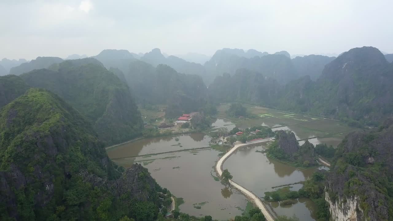 Cinematic drone footage of Tam Coc surrounded by majestic limestone peaks in misty and foggy weather. Ricefields in the wild jungle in the spring with boat tour canal in, Ninh Binh, Vietnam