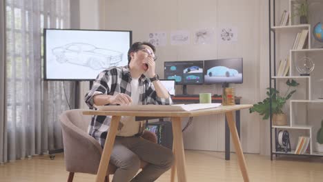 asian male yawning and sleeping while working on a car design sketch on table in the studio with tv and computers display 3d electric car model