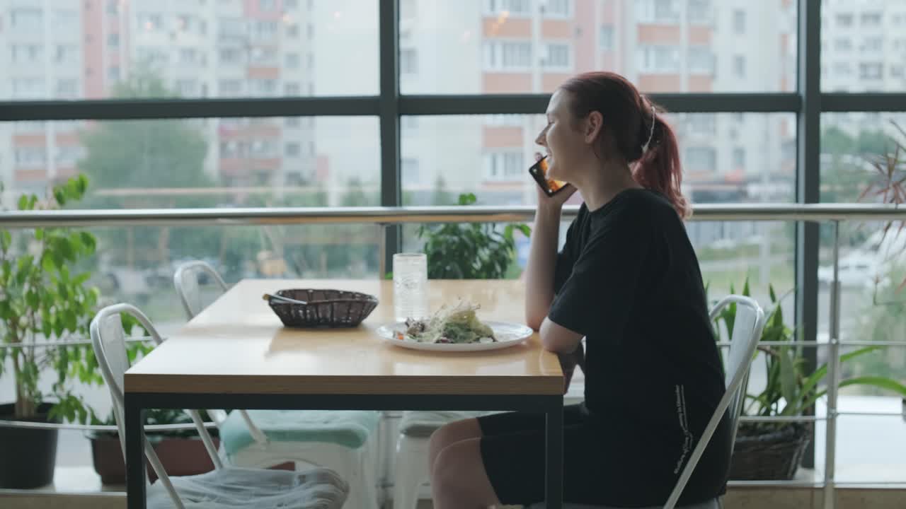 mujer joven comiendo una ensalada fresca en una mesa de madera en un restaurante moderno con grandes ventanas, luz natural y vistas urbanas. escena de comedor relajada