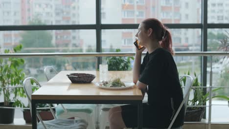 young woman eating a fresh salad at a wooden table in a modern restaurant with large windows, natural light, and urban views. relaxed dining scene