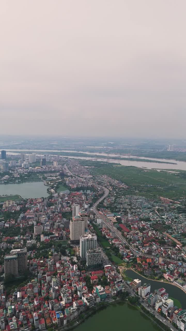 Wide vertical drone shot of Hanoi’s West Lake and Tay Ho district, blending water, modern buildings, and urban sprawl under soft daylight in Vietnam’s capital city