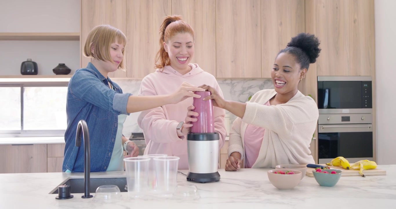 Diverse amiche preparano un frullato, posizionando una tazza rosa sulla base del frullatore sul bancone, stabilizzandola e allineandola.