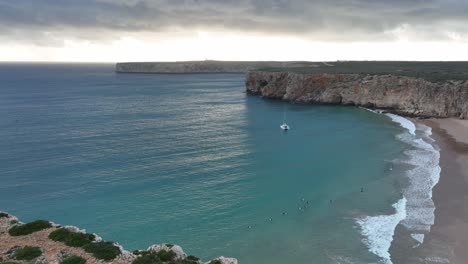 Spectacular-aerial-shot-of-an-anchored-catamaran-and-surfers-in-a-bay-of-turquoise-water-surrounded-by-cliffs
