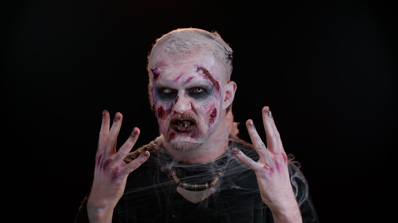 aterrador zombie herido no muerto tipo haciendo caras, mira a la cámara escondiéndose a través de las manos sonríe terriblemente