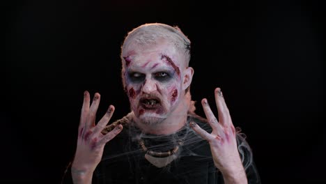 aterrador zombie herido no muerto tipo haciendo caras, mira a la cámara escondiéndose a través de las manos sonríe terriblemente