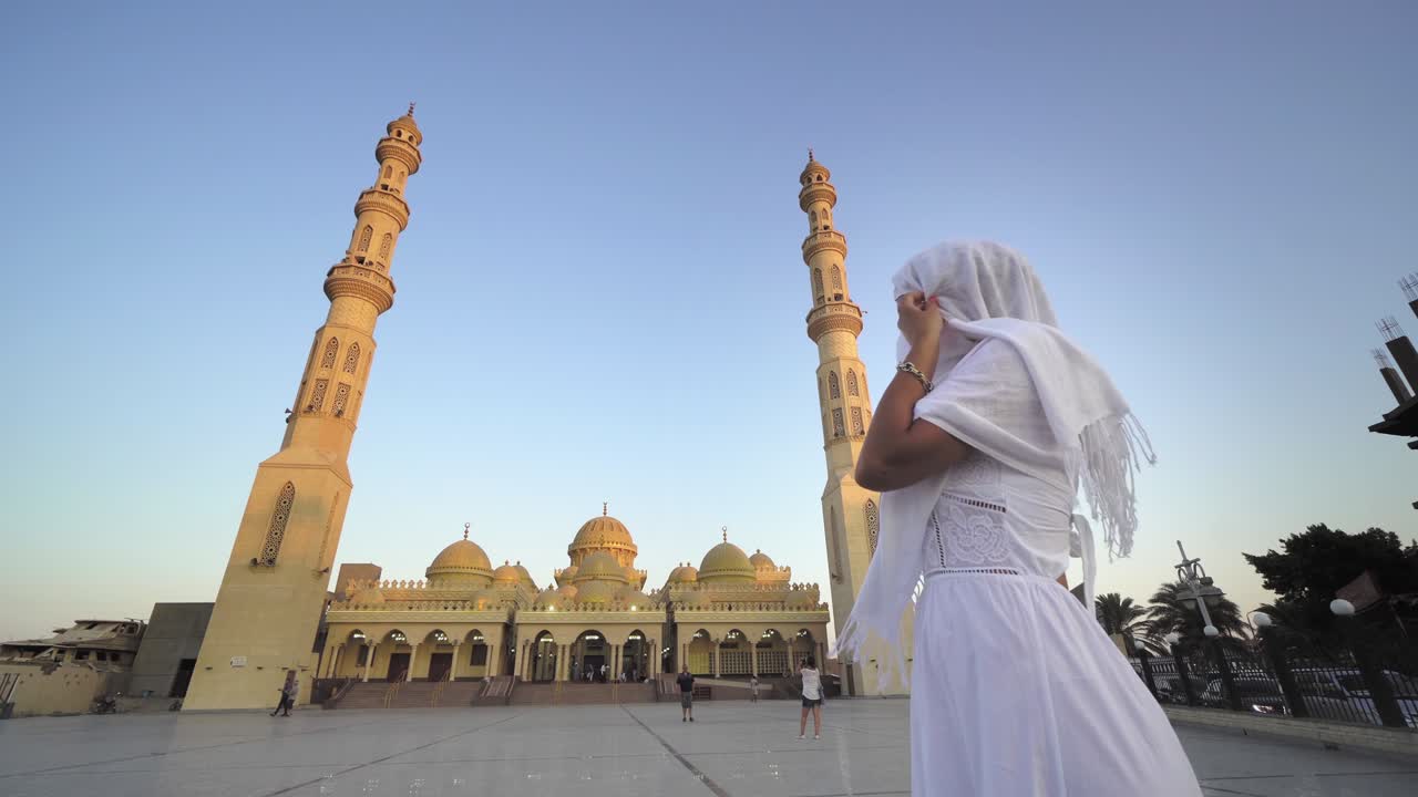 일몰과 해가 뜨는 엘 미나 모스크의 풍경과 함께 긴  드레스와 히자브를 입은 관광 여성이 모스크의 전망을 즐기고 있습니다.