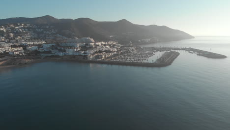 drone captures stunning footage of a coastal town port, with magnificent mountain range and charming housing estates nestled in background during early morning