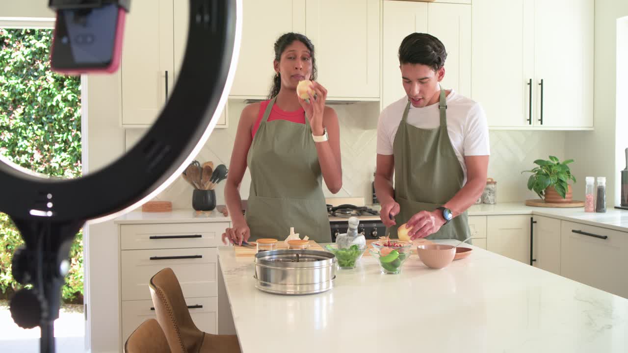 Casal afro-americano de aventais verdes filmando, preparando cebolas com ring light na ilha da cozinha